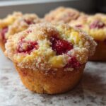 A close-up of freshly baked strawberry muffins topped with coarse sugar, showing bright red fruit inside.