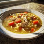 Close-up of a warm bowl of amazing turkey soup featuring shredded turkey, egg noodles, carrots, and celery in rich broth.