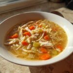 A white bowl filled with amazing turkey soup featuring shredded turkey, egg noodles, carrots, and celery, highlighted by sunlight.