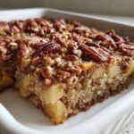 A thick slice of apple pecan baked oatmeal with a crunchy topping, sitting in a white baking dish.