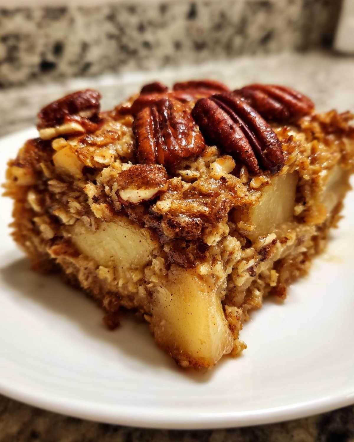 Close-up of a square slice of apple baked oatmeal topped with pecans, served on a white plate.