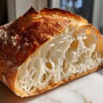 Close-up of a freshly baked artisan bread loaf cut in half, showing an open, airy crumb structure and a deeply browned, blistered crust.