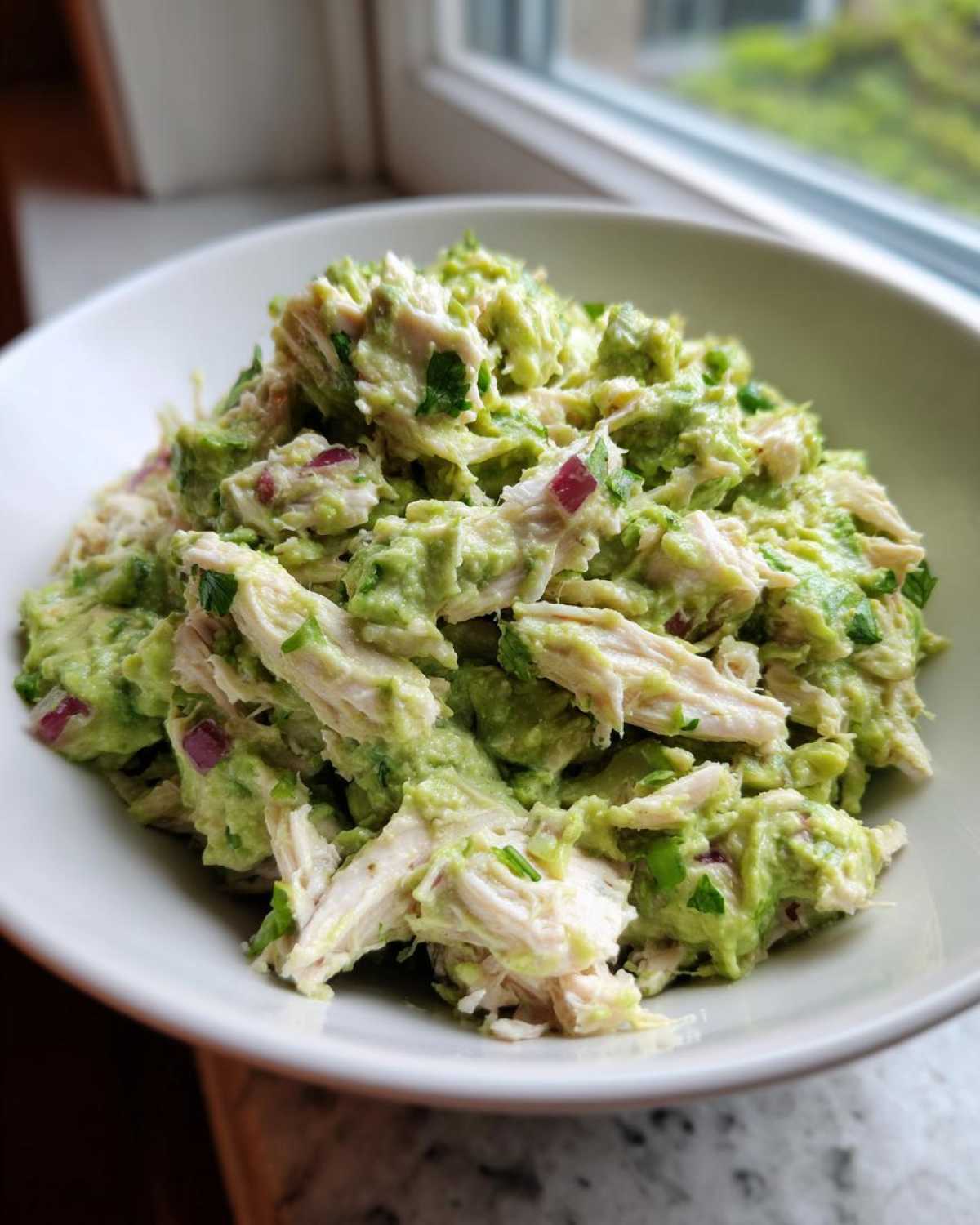 Close-up of creamy avocado chicken salad with shredded chicken and red onion pieces in a white bowl.