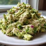 A generous mound of creamy avocado chicken salad mixed with shredded chicken, red onion, and cilantro on a white plate.