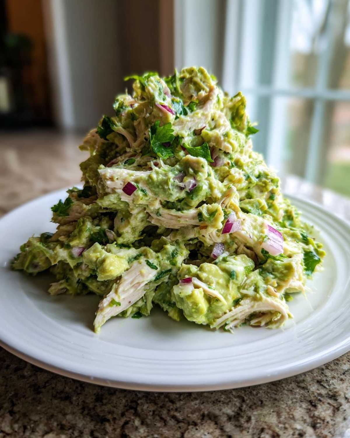 A mound of creamy avocado chicken salad mixed with shredded chicken and diced red onion, garnished with parsley.