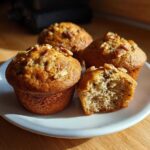 Four golden brown banana nut muffins on a white plate, one is broken open showing the moist crumb.