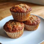 Three golden-brown banana protein muffins stacked on a white plate against a wooden background.