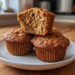 A stack of three golden-brown banana protein muffins, one cut open to show the moist interior.