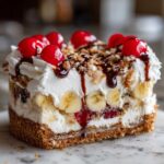 A close-up view of a decadent slice of banana split cake featuring layers of cream, bananas, and topped with cherries and chocolate sauce.