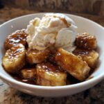 A white bowl filled with caramelized bananas foster and a scoop of vanilla ice cream melting on top.