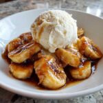 A white bowl filled with caramelized bananas in rich sauce, topped with a scoop of vanilla ice cream, making up the perfect bananas foster.