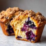 A close-up of a perfectly baked blueberry muffin, cut in half to show the moist interior packed with blueberries and a sugary crumb topping.