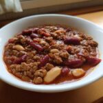 A close-up of a white bowl filled with the best chili recipe, featuring ground meat, kidney beans, and white beans in a rich red sauce.