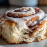 A close-up, mouth-watering shot of a single, soft cinnamon roll topped generously with white cream cheese frosting.
