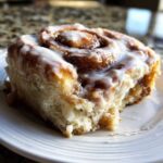 A close-up, appetizing photo of a single, fluffy cinnamon roll topped generously with white icing.