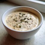 Close-up of a white bowl filled with creamy, light-colored cream of chicken soup, garnished with fresh thyme sprigs and cracked black pepper.