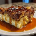 A close-up of a square slice of bread pudding with bourbon sauce generously drizzled over the top and pooled on the white plate.