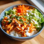 A close-up of a vibrant buffalo chicken bowl featuring shredded buffalo chicken over white rice, topped with ranch and green onions.