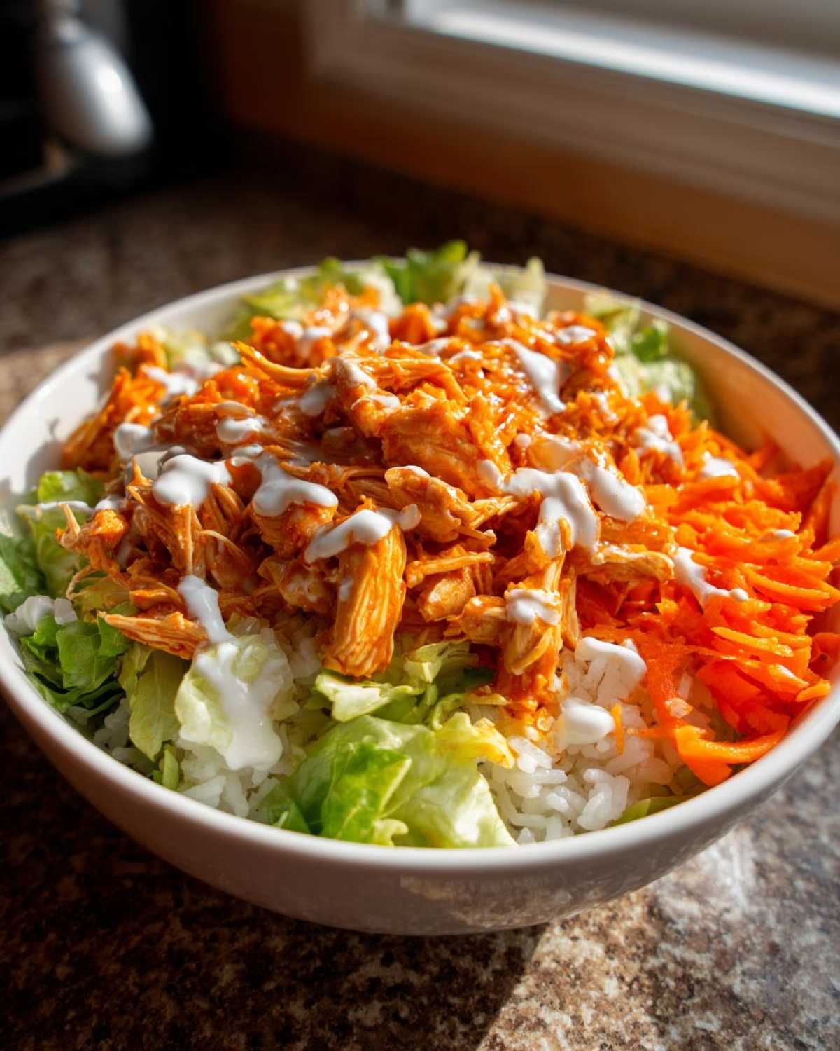 A close-up of a vibrant buffalo chicken bowl featuring shredded buffalo chicken, lettuce, rice, carrots, and a ranch drizzle.
