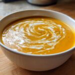 A close-up of vibrant orange butternut squash soup in a white bowl, garnished with a swirl of cream and black pepper.