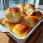 A close-up of golden brown, buttery dinner rolls stacked in a white baking dish, glistening under natural light.