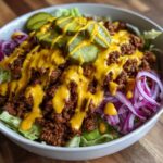 A close-up of a delicious cheeseburger bowl featuring seasoned ground beef, lettuce, red onion, pickles, and a drizzle of cheese sauce.