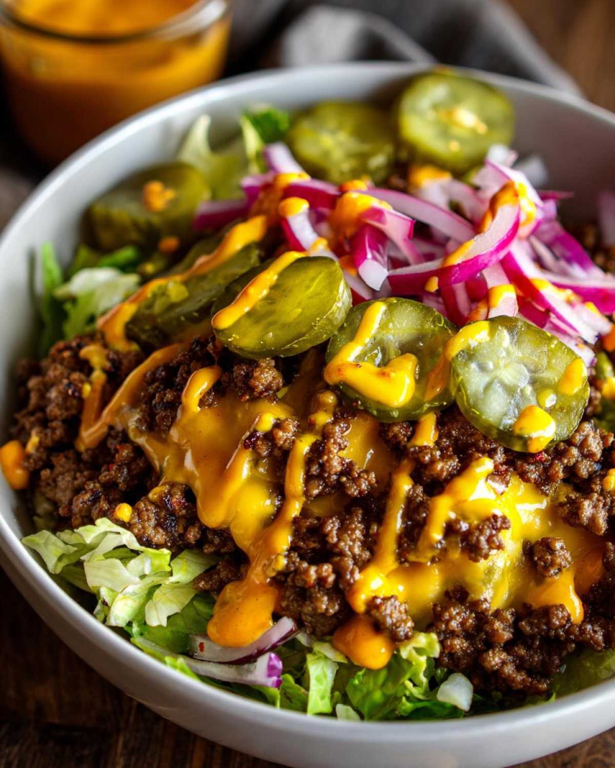 Close-up of a fresh cheeseburger bowl featuring seasoned ground beef, melted cheese sauce, pickles, and red onion over lettuce.
