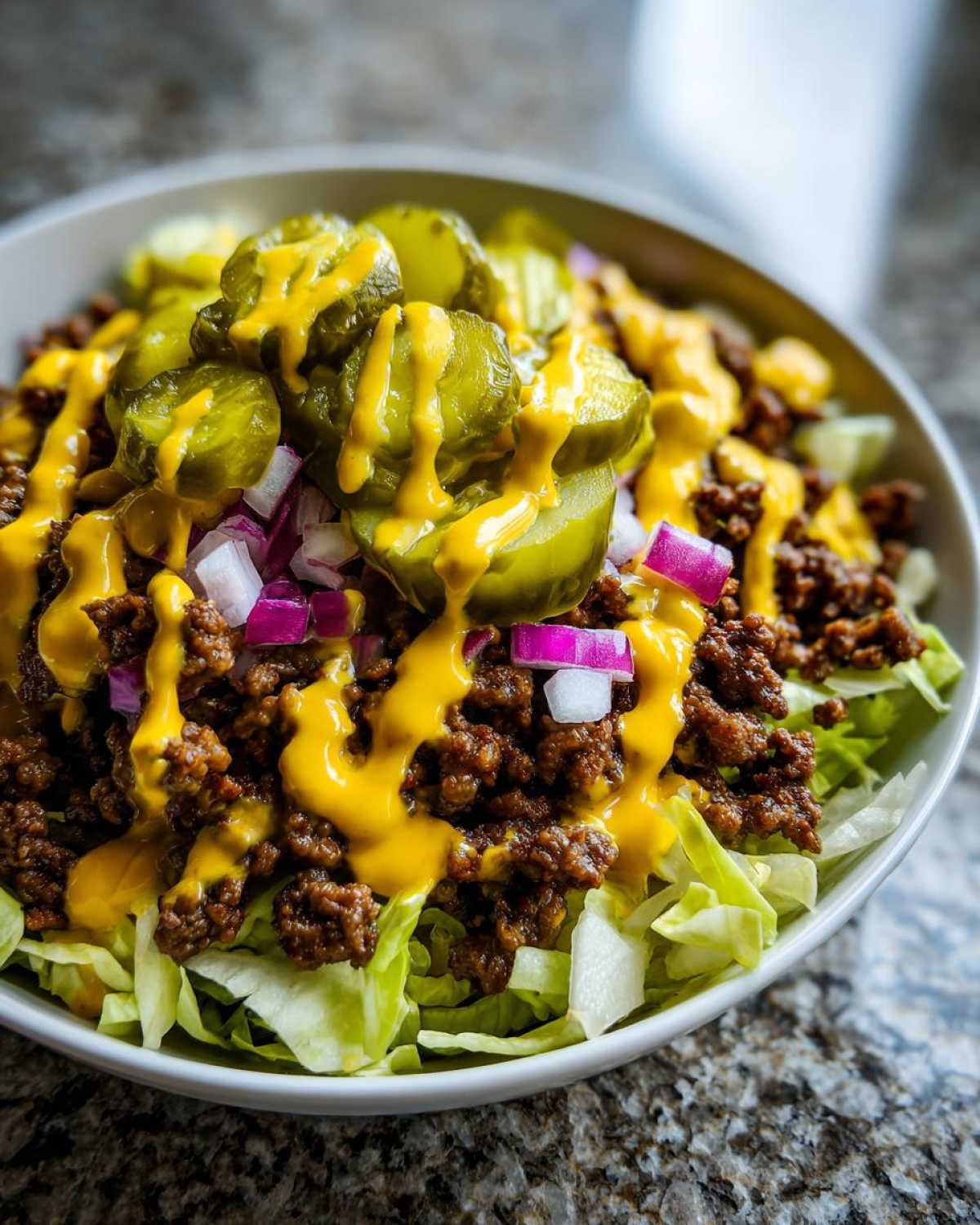 A close-up of a delicious cheeseburger bowl featuring seasoned ground beef over lettuce, topped with pickles, red onion, and a drizzle of cheese sauce.