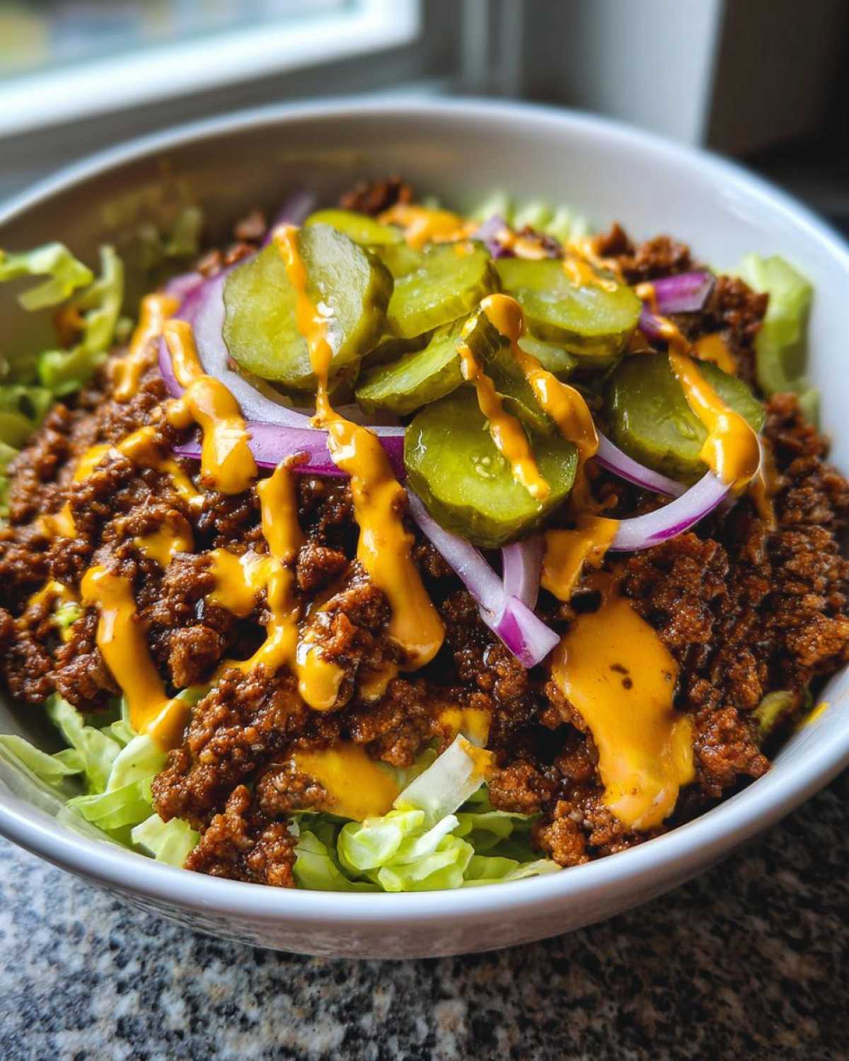 A close-up of a cheeseburger bowl featuring seasoned ground beef, shredded lettuce, red onion, pickles, and a drizzle of cheese sauce.