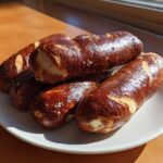 A stack of freshly baked, dark brown, chewy pretzel dogs sprinkled with coarse salt on a white plate.