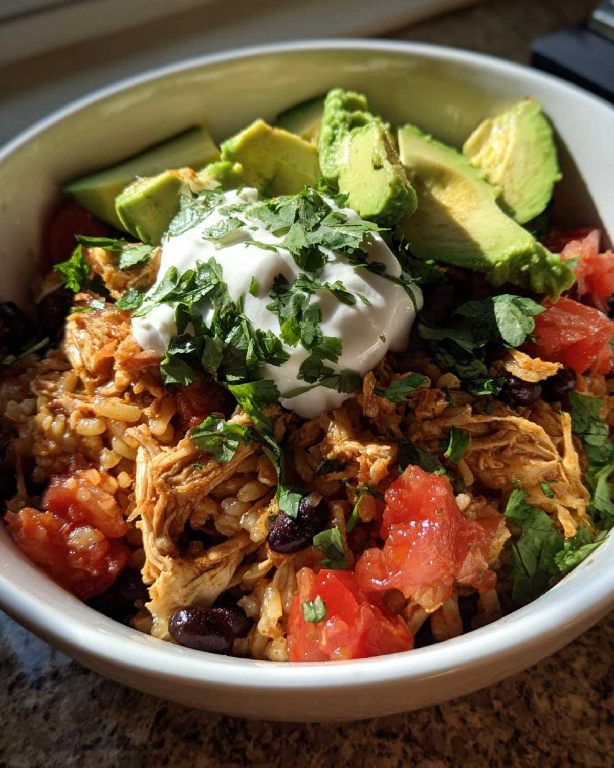 A close-up of a delicious chicken burrito bowl topped with shredded chicken, rice, black beans, avocado, and sour cream.