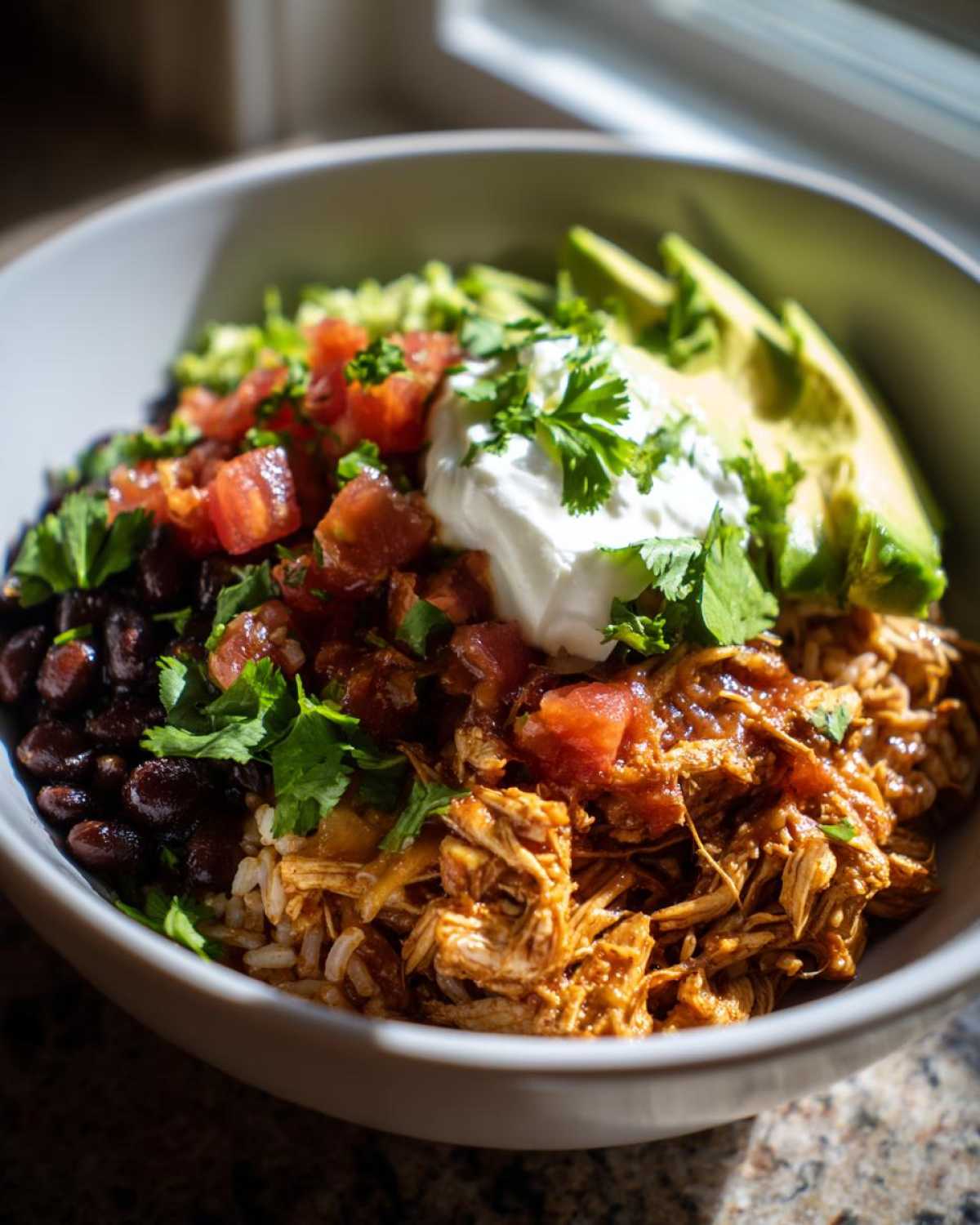 Close-up of a vibrant chicken burrito bowl featuring shredded seasoned chicken, black beans, rice, salsa, sour cream, and avocado slices.