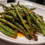 A close-up of blistered, bright green chinese green beans with garlic sauce served on a white plate.
