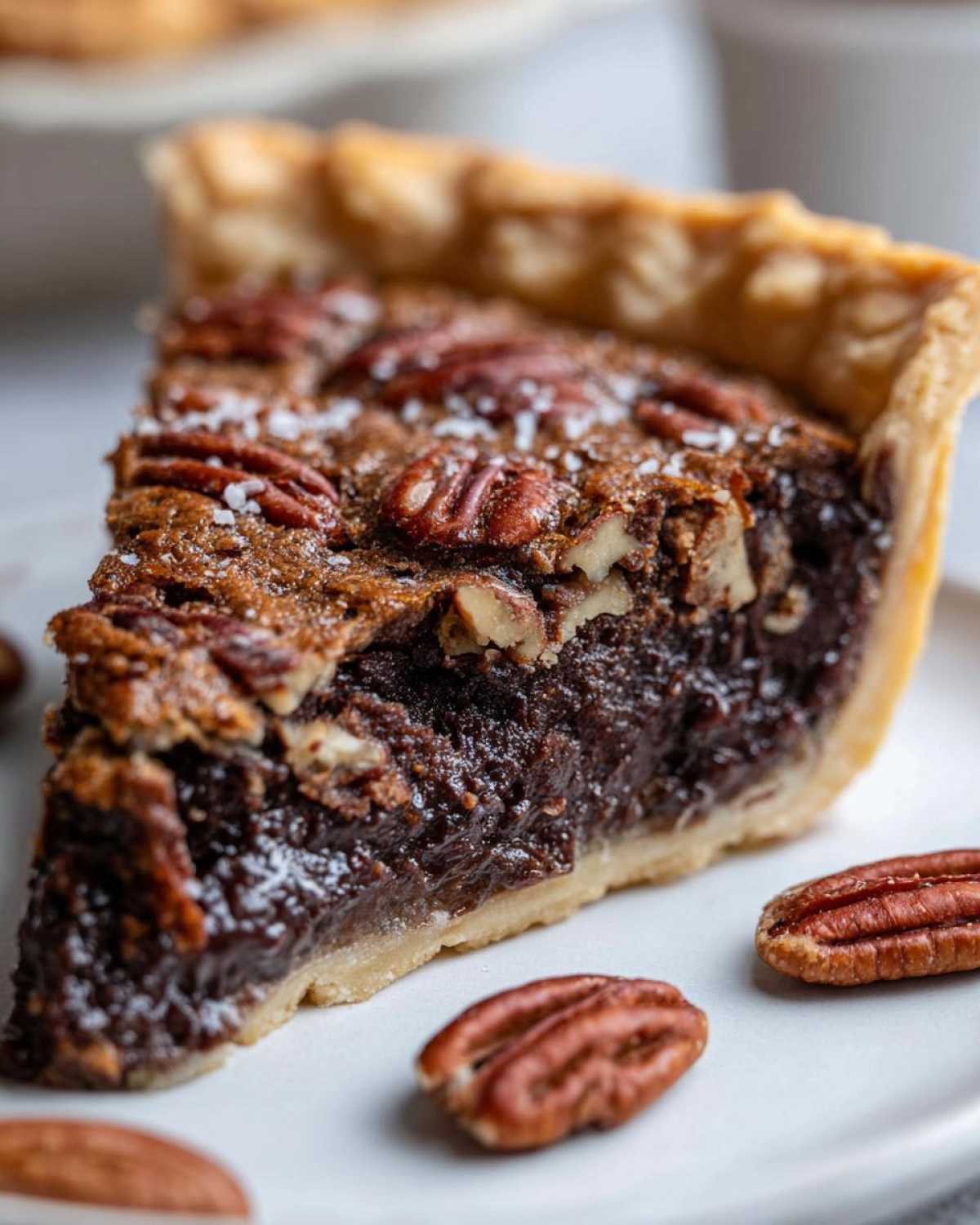 A close-up slice of decadent chocolate pecan pie topped with pecans and flaky sea salt.