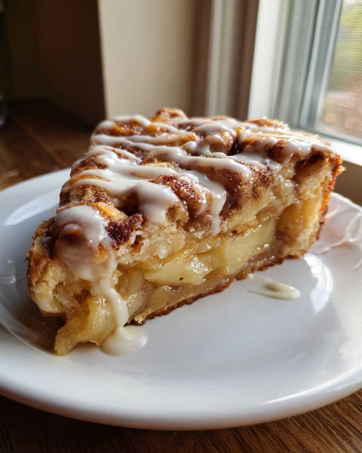 A close-up slice of cinnamon roll apple pie, showing layers of cooked apples and cinnamon swirl, topped with white icing.
