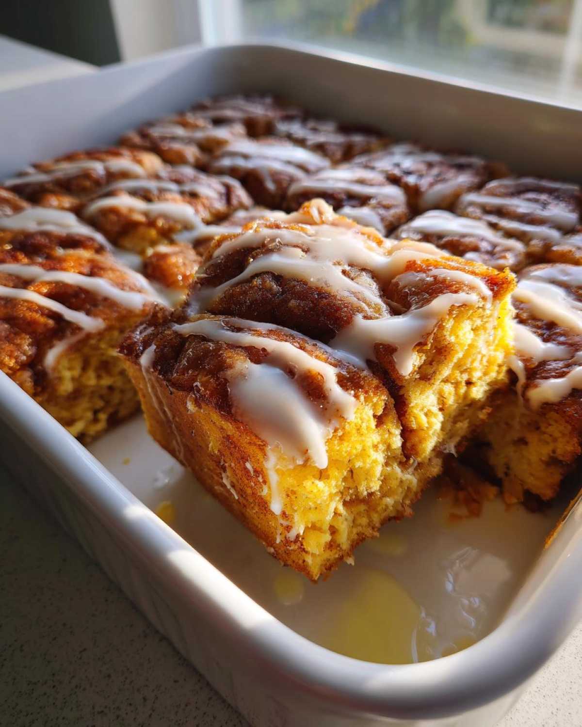 A close-up of a square portion of warm Cinnamon roll casserole drizzled with white icing in a white baking dish.