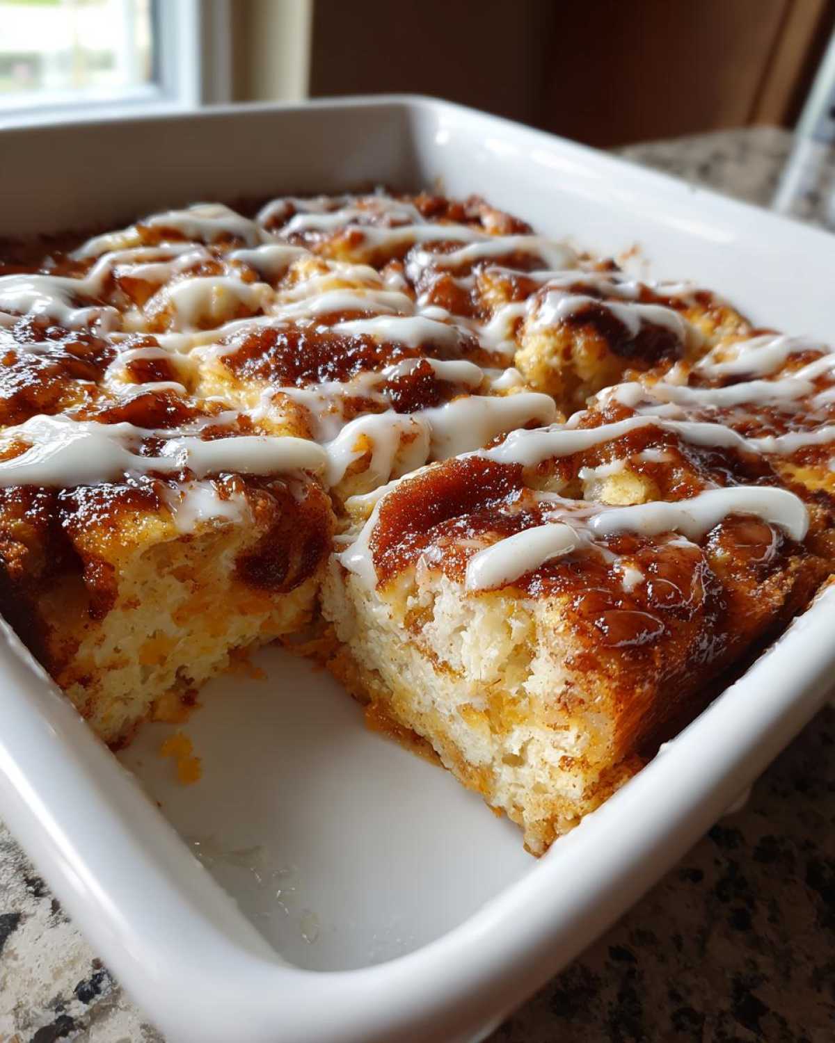 A close-up of a freshly baked cinnamon roll casserole in a white baking dish, drizzled with white icing.