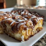 A close-up, low-angle shot of a freshly baked slice of cinnamon roll casserole topped with white icing.