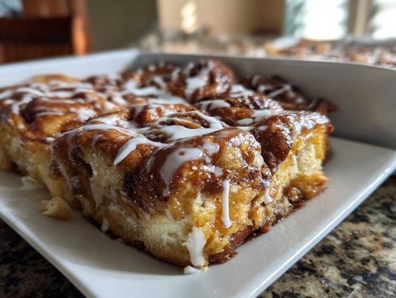 A close-up, low-angle shot of a freshly baked slice of cinnamon roll casserole topped with white icing.