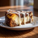 A rich slice of cinnamon roll cheesecake topped with thick white icing drizzle, sitting on a white plate.