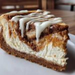 A close-up of a rich slice of cinnamon roll cheesecake with a thick graham cracker crust and white icing drizzled on top.