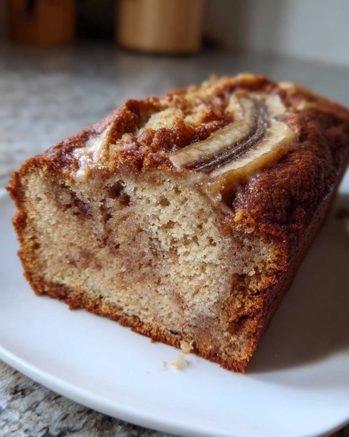 A close-up of a slice of moist cream cheese banana bread with a caramelized banana slice on top, served on a white plate.