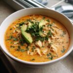 A close-up of a white bowl filled with creamy chicken tortilla soup, topped with shredded chicken, avocado slices, and fresh cilantro.