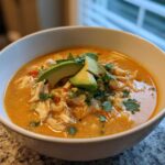 A close-up of a white bowl filled with creamy chicken tortilla soup, topped with shredded chicken, avocado slices, and cilantro.