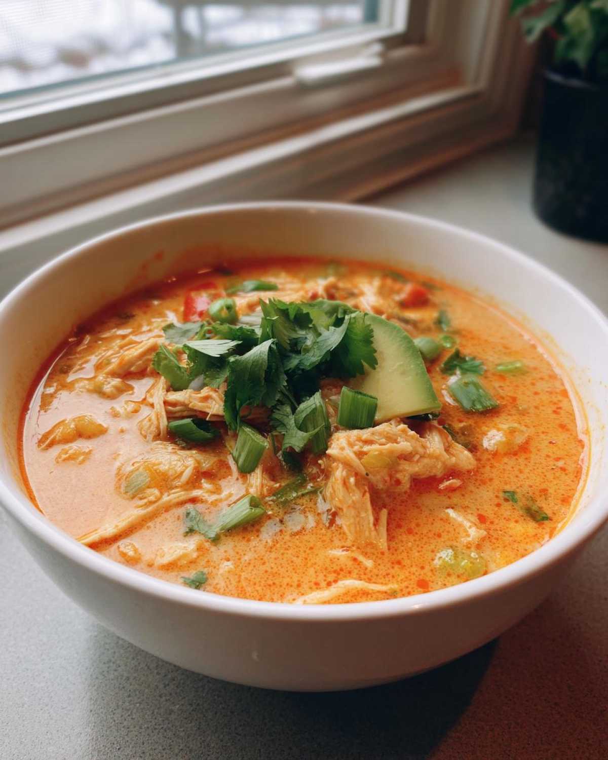 A white bowl filled with creamy chicken tortilla soup, topped with shredded chicken, avocado, and fresh cilantro.