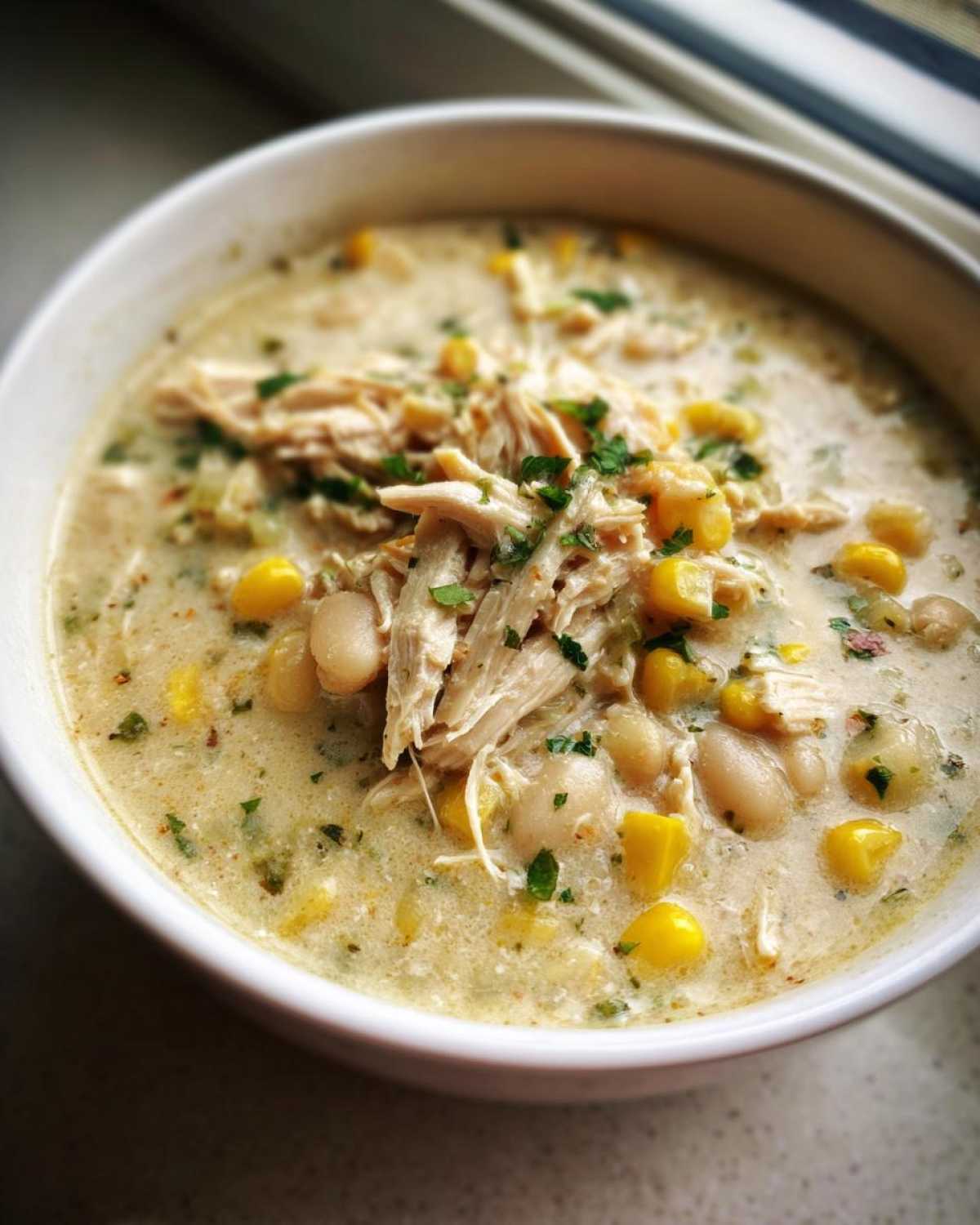 Close-up of a white bowl filled with creamy crockpot white chicken chili, topped with shredded chicken, white beans, corn, and parsley.