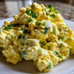 A generous scoop of creamy egg salad recipe garnished with fresh chopped green onions on a white plate.