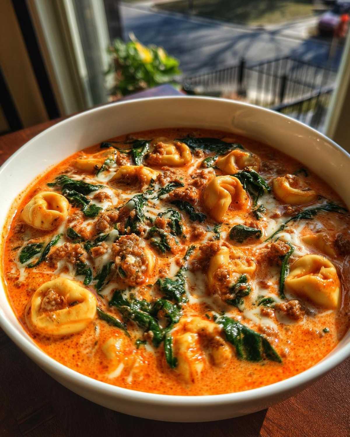 Close-up of a bowl of creamy italian sausage tortellini soup with spinach, garnished with a swirl of cream.