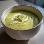 Close-up of a creamy, pale green bowl of leek soup, swirled with cream and topped with fresh herbs.