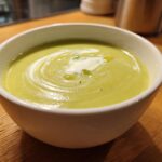 A close-up of a white bowl filled with smooth, light green leek soup, garnished with a swirl of cream and olive oil.